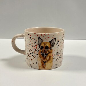 A ceramic mug with a painted German Shepherd and speckles of red and black paint against a white background.