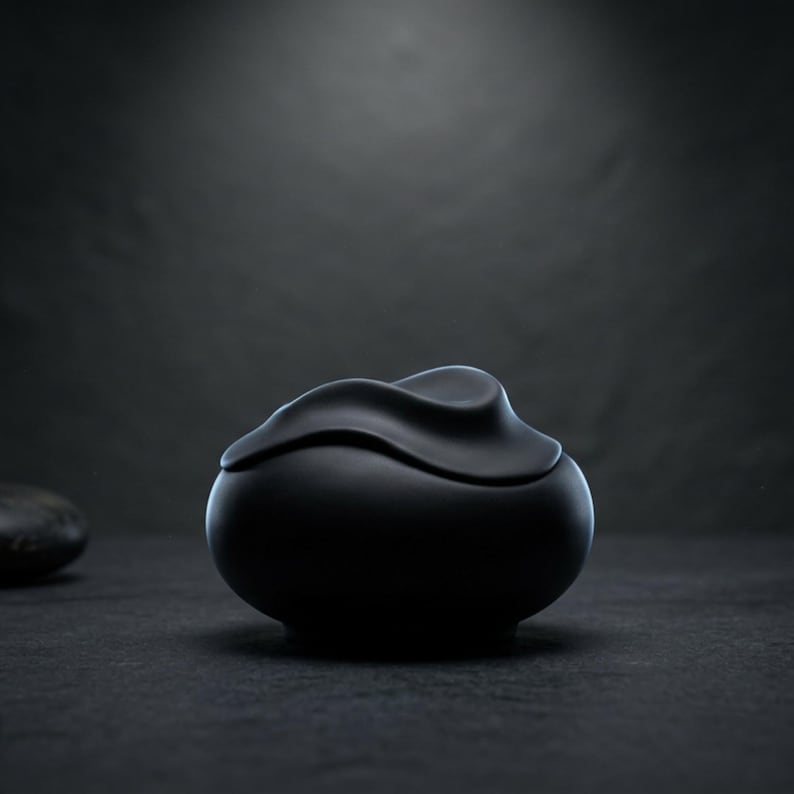 Pode incluir: Um recipiente de cer&acirc;mica preta fosca com tampa. A tampa tem uma forma ondulada e org&acirc;nica. O recipiente est&aacute; sobre uma superf&iacute;cie escura, com um fundo escuro desfocado. O design &eacute; minimalista e moderno.