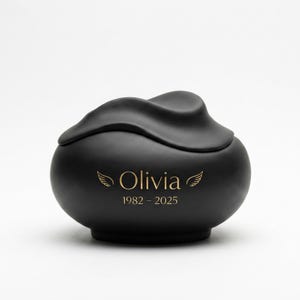 Pode incluir: Uma urna preta fosca com um design de tampa ondulada. A urna est&aacute; inscrita com o nome "Olivia", motivos de asas e os anos "1982 - 2025" em dourado. A urna tem uma forma arredondada.