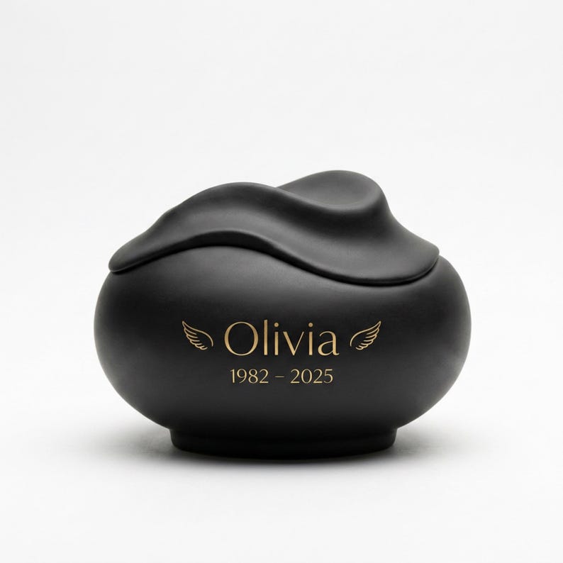 Pode incluir: Uma urna preta fosca com um design de tampa ondulada. A urna est&aacute; inscrita com o nome "Olivia", motivos de asas e os anos "1982 - 2025" em dourado. A urna tem uma forma arredondada.