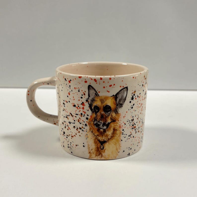 A ceramic coffee mug painted with a German Shepherd and colorful splatters stands on a white surface.
