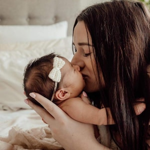 May include: A mother kisses her baby, a tender moment captured in a soft, natural light. The baby wears a white bow headband. The scene evokes a sense of warmth and love, with the focus on the intimate connection.