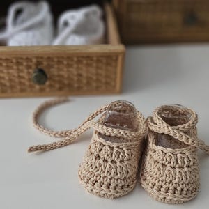 May include: A pair of beige crochet baby shoes featuring a rounded toe, a low-cut opening, and long matching crochet laces that wrap around the ankle. The shoes are crafted with a textured stitch pattern and are positioned on a flat surface.