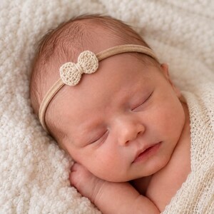 May include: A thin, beige elastic headband featuring a small, centred, crocheted bow in a matching light tan colour. The accessory is designed with a simple, minimalist construction, showcasing a delicate texture on the bow and a smooth band.