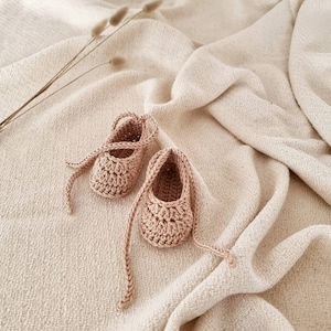 May include: A pair of light beige crochet baby booties featuring a textured stitch pattern and long, braided ties that wrap around the ankle. The footwear is crafted from a soft, neutral-toned yarn, showcasing a rounded toe design and an open top.