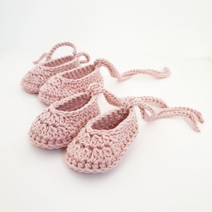 May include: Two pairs of dusty pink crochet baby booties featuring a textured stitch pattern, a rounded toe design, and long matching yarn ties attached to the sides for securing the footwear around the ankle area in a simple bow configuration.
