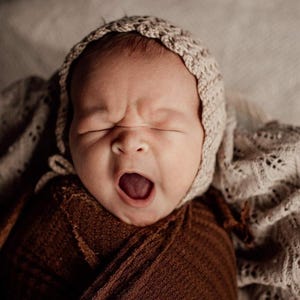 May include: A newborn baby yawning, wearing a beige knitted bonnet and wrapped in a brown blanket. The baby's mouth is wide open, and its eyes are closed. The background includes a soft, patterned blanket.