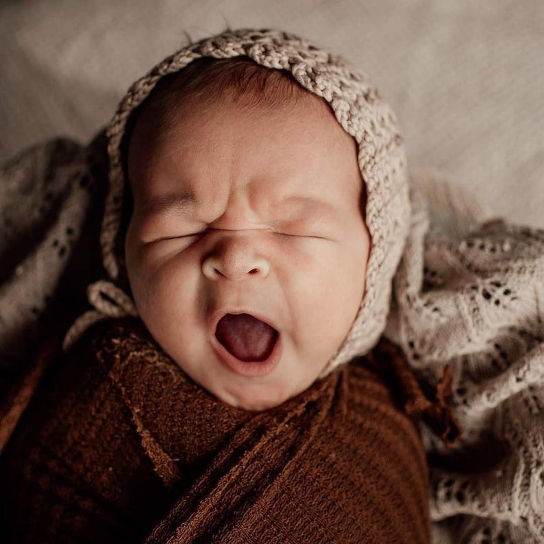 May include: A newborn baby yawning, wearing a beige knitted bonnet and wrapped in a brown blanket. The baby's mouth is wide open, and its eyes are closed. The background includes a soft, patterned blanket.