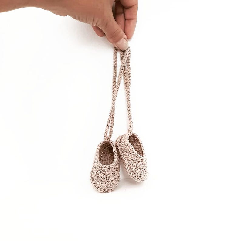 May include: A pair of small, light beige crochet baby booties connected by a long, thin matching yarn cord. The footwear features a textured, woven stitch pattern with a rounded toe and an open top, crafted from a neutral-colored yarn material.