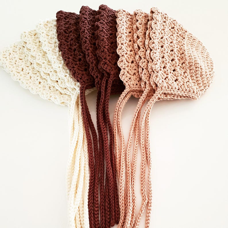 May include: A collection of five crocheted baby bonnets in various colors: cream, deep burgundy, medium burgundy, light peach, and peach. Each bonnet features a scalloped edge and long, braided ties for securing under the chin. The bonnets are arranged in a row.
