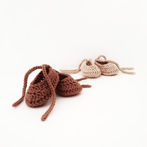 May include: Two pairs of handmade crochet baby booties with ankle ties. One pair is dark brown and the other pair is light beige. The booties feature a textured yarn construction with a rounded toe design and long braided straps for securing.