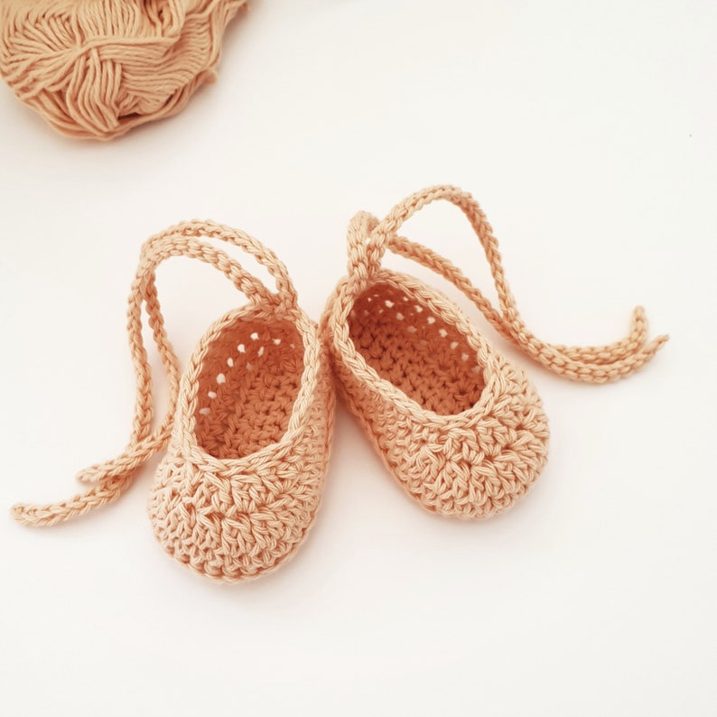 May include: A pair of peach-colored crochet baby booties featuring a textured stitch pattern, a rounded toe, and long matching yarn ties attached to the heel area for securing the footwear, accompanied by a ball of matching yarn.