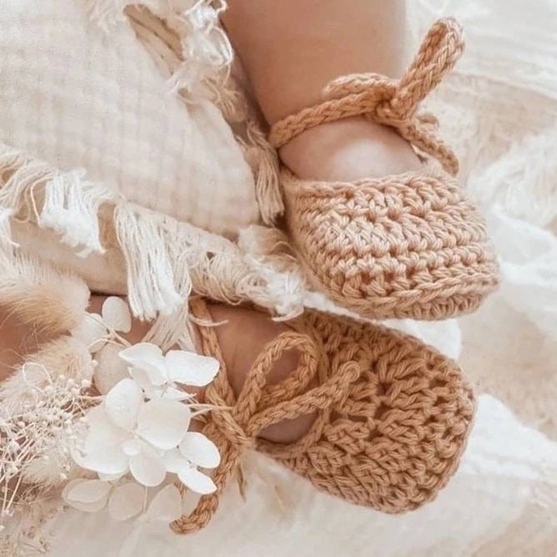 May include: A pair of tan crochet baby booties featuring a rounded toe, a woven texture, and ankle straps that tie into a bow. One bootie is adorned with a cluster of white dried flowers and small white petals attached to the side strap.