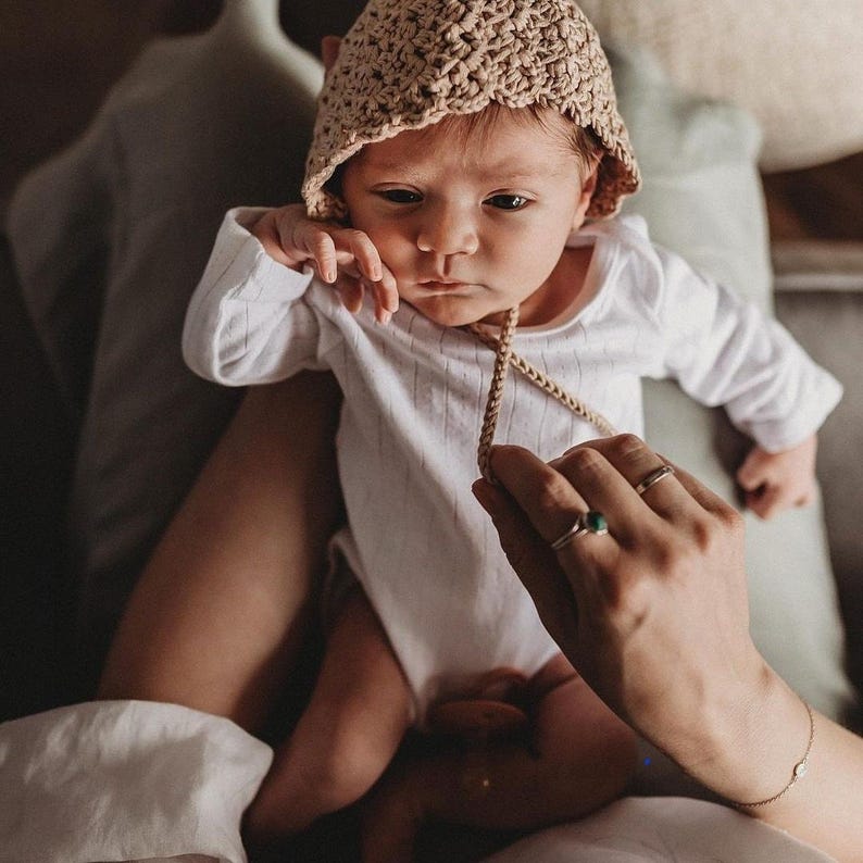 May include: A newborn baby wearing a beige crocheted bonnet and a white ribbed onesie. The baby is being held, with a hand visible holding the bonnet's tie. The baby's expression is serious.