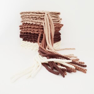 May include: A stack of crocheted items in a gradient of neutral colors, from cream to dark brown. The stack has a textured, scalloped edge. Long, braided strands of yarn in matching colors cascade from the top, creating a layered, handmade aesthetic.