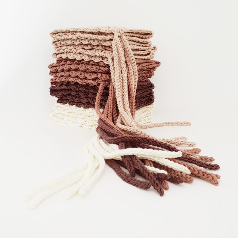 May include: A stack of crocheted items in a gradient of neutral colors, from cream to dark brown. The stack has a textured, scalloped edge. Long, braided strands of yarn in matching colors cascade from the top, creating a layered, handmade aesthetic.