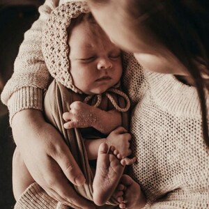 May include: A newborn baby wearing a beige bonnet and wrapped in a matching blanket is held in someone's arms. The baby's eyes are closed, and the hands and feet are visible. The person holding the baby is wearing a cream-colored sweater.