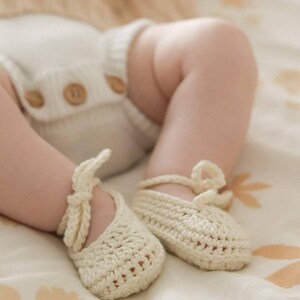 May include: A pair of cream-colored crocheted baby booties with ankle ties, paired with a matching cream-colored ribbed knit diaper cover featuring three brown wooden buttons at the front waistband, worn on a pair of infant legs.