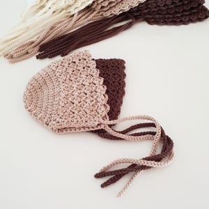 May include: Several handmade crochet bonnets in various shades of brown and cream. The bonnets have a scalloped edge and long ties. The crochet work appears to be tight and even, suggesting a high-quality product. The bonnets are likely for babies or young children.