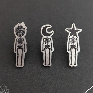 May include: Three silver skeleton pins with different head designs. The first pin has a flame design, the second has a crescent moon design, and the third has a star design.