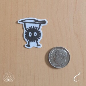May include: A black and white sticker depicting a round, spiky character with two eyes and two legs, holding a knife above its head. The sticker is placed next to a silver United States dime coin, which provides a clear indication of the item's scale.