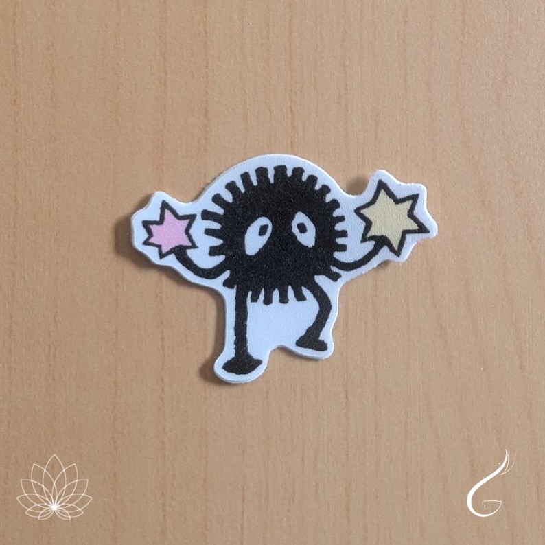 May include: A die-cut sticker featuring a black, spiky, circular creature with two large white eyes. The creature has thin arms and legs, and is holding a small, light pink star in its left hand and a pale yellow star in its right hand.