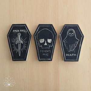 May include: Three black wooden coffin-shaped boxes with white text and illustrations. The first box says "Amor Fati" and has a sword illustration. The second box says "Memento Mori" and has a skull illustration. The third box says "Death" and has a ghost illustration.