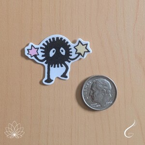 May include: A die-cut sticker featuring a black, spiky, circular creature with two white eyes, two thin arms holding a pink star and a yellow star, and two legs. A silver-coloured United States dime is placed next to the sticker for scale.