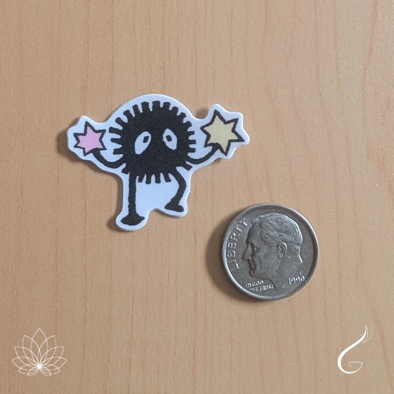 May include: A die-cut sticker featuring a black, spiky, circular creature with two white eyes, two thin arms holding a pink star and a yellow star, and two legs. A silver-coloured United States dime is placed next to the sticker for scale.