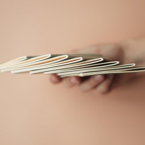 May include: A stack of blank, cream-colored notebooks held in a hand.