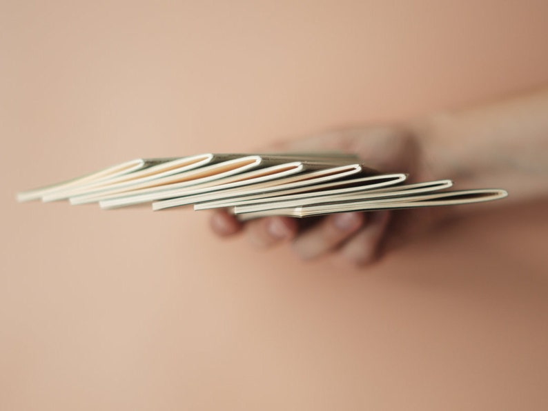 May include: A stack of blank, cream-colored notebooks held in a hand.