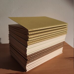 May include: A stack of notebooks with brown, beige, and green covers. The notebooks are bound with a stitched spine and have a subtle textured finish.