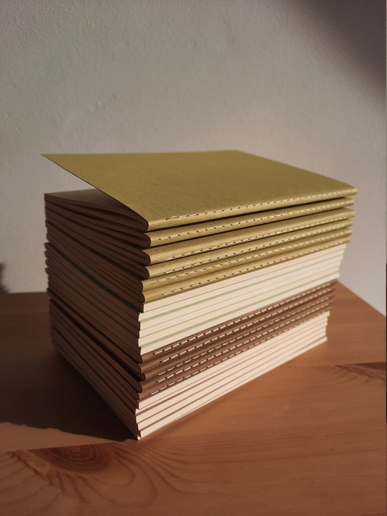 May include: A stack of notebooks with brown, beige, and green covers. The notebooks are bound with a stitched spine and have a subtle textured finish.