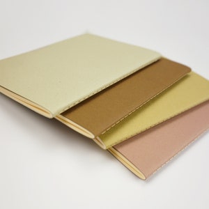 May include: Four small notebooks stacked on top of each other. The notebooks are different colors: beige, brown, yellow, and pink. The notebooks have a stitched binding.