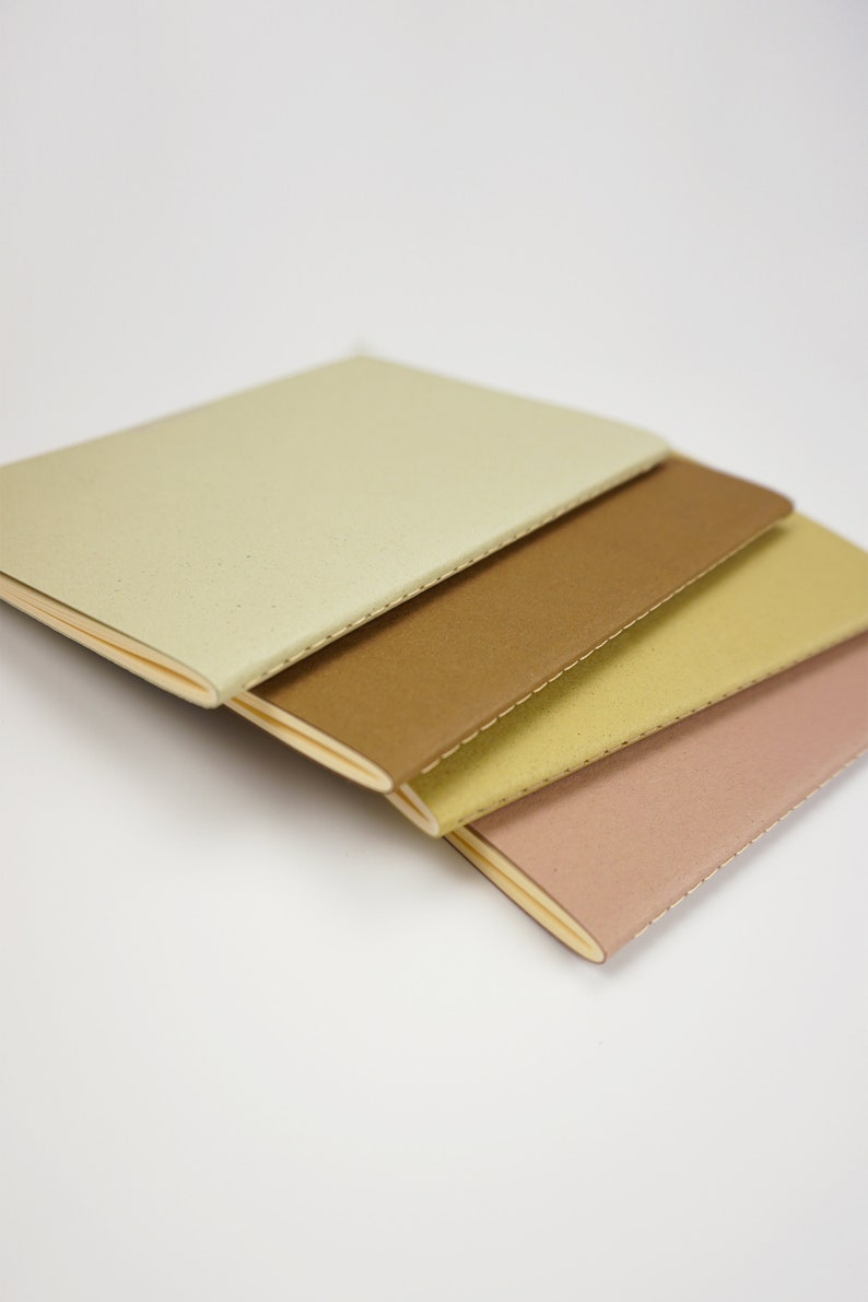 May include: Four small notebooks stacked on top of each other. The notebooks are different colors: beige, brown, yellow, and pink. The notebooks have a stitched binding.