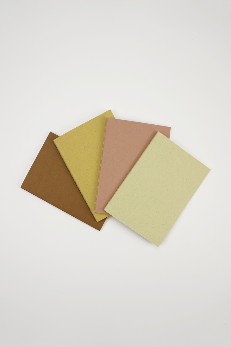 May include: Four small notebooks with different colored covers: brown, yellow, pink, and light green. The notebooks are arranged in a fan shape.