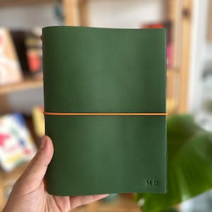 May include: A dark green leather notebook cover featuring a smooth matte finish and a bright orange elastic band wrapped horizontally around the centre. The bottom right corner is embossed with the capital letters MB in a simple sans-serif font.