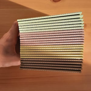 May include: A stack of 15 small, bound notebooks with stitched spines. The notebooks have covers in shades of brown, beige, and green.