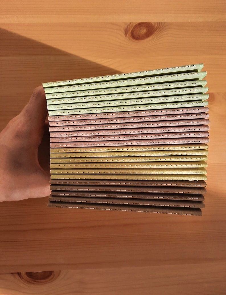 May include: A stack of 15 small, bound notebooks with stitched spines. The notebooks have covers in shades of brown, beige, and green.