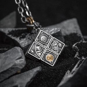 May include: A silver-toned pendant necklace with a geometric design. The pendant features four square panels with intricate patterns and a gold-colored accent. The necklace chain is also silver-toned. The pendant is approximately 1.5 inches square.