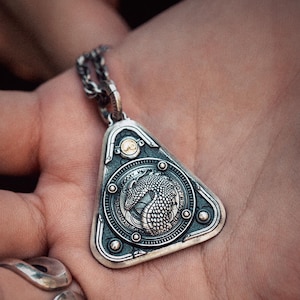 Könnte beinhalten: Silberne Anhänger-Halskette mit dreieckigem Rahmen und rundem Mittelteil mit detailliertem Drachendesign. Der Anhänger hat eine strukturierte, antike Oberfläche und hängt an einer Silberkette.