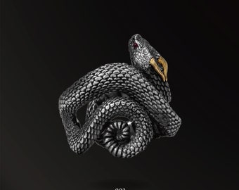 May include: A silver ring with a detailed snake design. The snake is coiled and has a red eye and gold accents. The ring is labeled "002 HORN SNAKES".