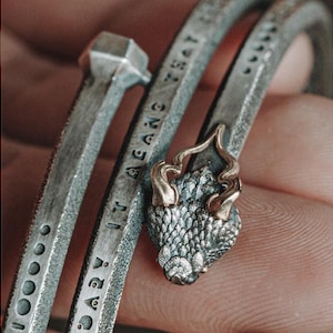 Könnte beinhalten: Nahaufnahme von drei silbernen Metallarmbändern. Eines hat ein Nagelkopfdessign, ein anderes den Text "BABY IT MEANS THAT" eingraviert, und das dritte zeigt einen Drachenkopf mit goldenen Akzenten.