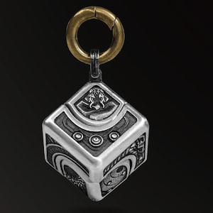Könnte beinhalten: Ein silberner Anhänger mit einem geometrischen Würfeldesign, das komplizierte Muster und einen goldfarbenen Ring an der Oberseite aufweist. Der Anhänger hat eine detaillierte, geprägte Oberfläche mit kreisförmigen und geschwungenen Elementen.