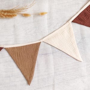 May include: A decorative banner with triangular flags in shades of brown and cream, trimmed with a cream-colored border. The banner is made of textured fabric and is suitable for home decor.
