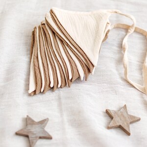 May include: A stack of beige and brown fabric triangle flags with a cream-colored ribbon, and three wooden star decorations. The flags are layered, creating a textured effect. The stars are scattered around the flags on a white fabric surface.
