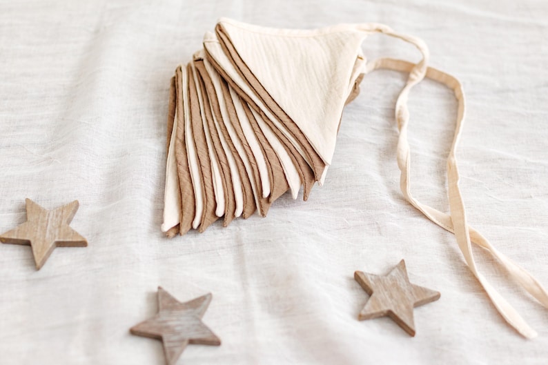May include: A stack of beige and brown fabric triangle flags with a cream-colored ribbon, and three wooden star decorations. The flags are layered, creating a textured effect. The stars are scattered around the flags on a white fabric surface.
