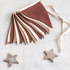 May include: A decorative banner with triangular flags in shades of brown, beige, and cream, strung together with a cream-colored ribbon. Three wooden star ornaments are scattered around the banner on a white surface.