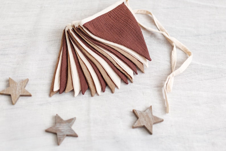 May include: A decorative banner with triangular flags in shades of brown, beige, and cream, strung together with a cream-colored ribbon. Three wooden star ornaments are scattered around the banner on a white surface.