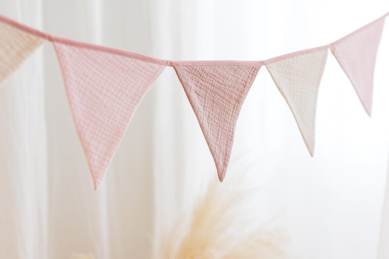 May include: A pink and white fabric bunting with triangular flags. The flags are made of a soft, gauzy material and are strung together with a white cord.